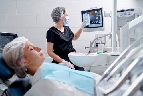 Patient receiving dental treatment with monitoring equipment while a dental professional supervises sedation.