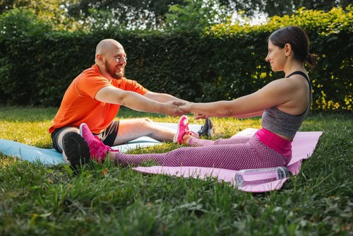 A man and woman supporting each other during an outdoor stretching session, demonstrating how accountability partners can encourage consistency, motivation and progress towards fitness goals.